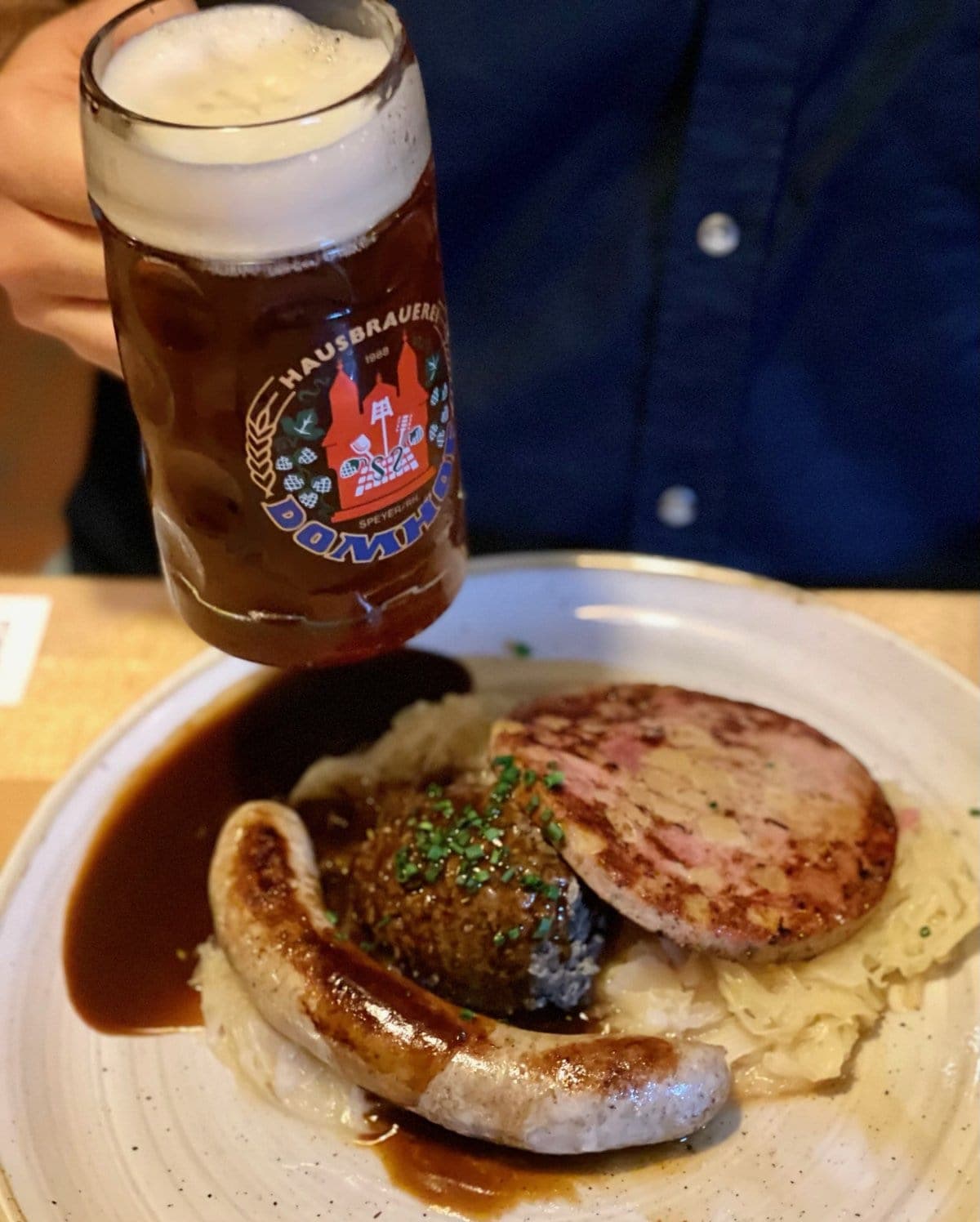 A person holding a glass of dark beer with the logo "Hausschraenke," paired with traditional German sausages, mashed potatoes, and gravy on a dinner plate.