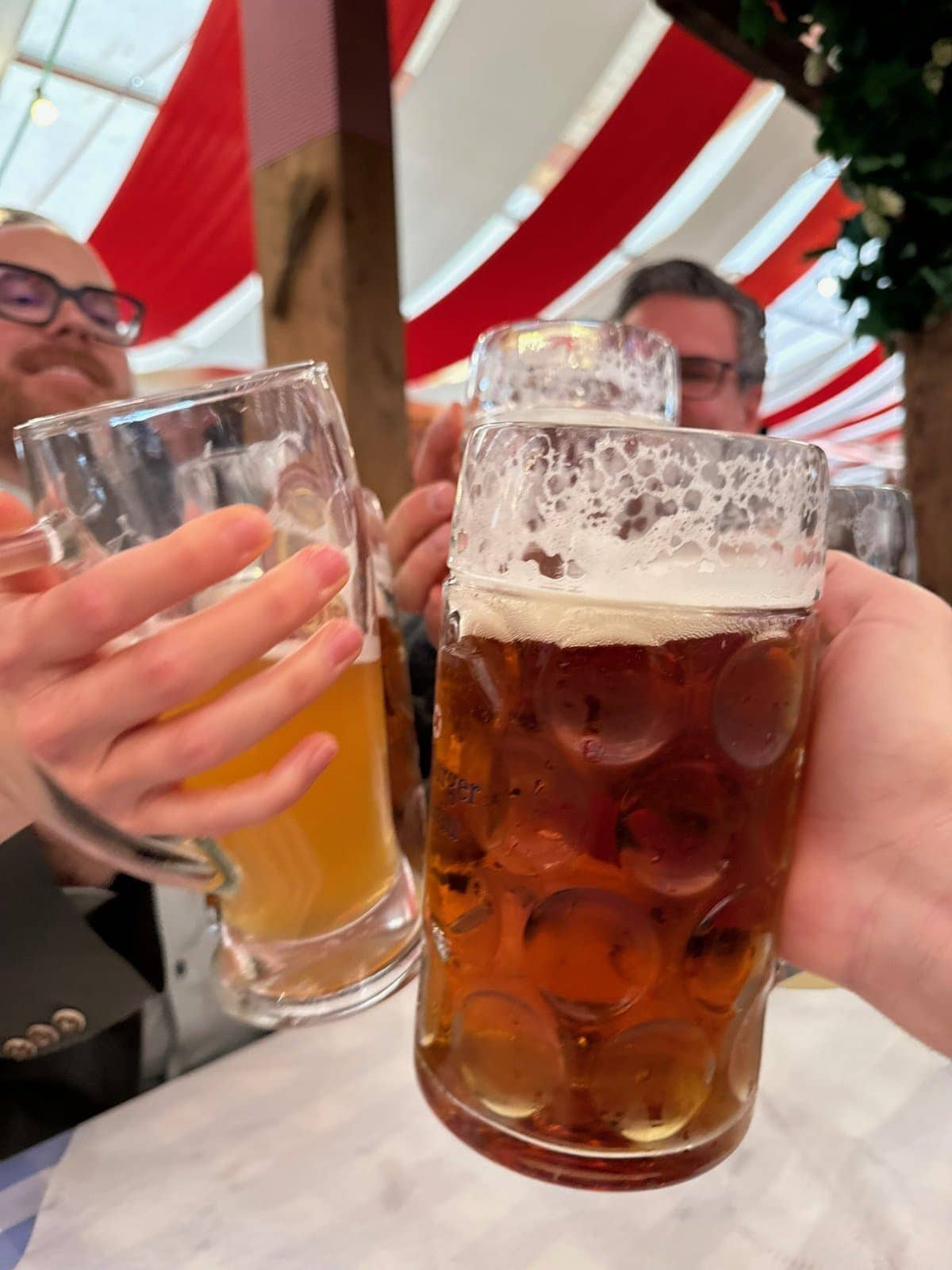Beer mugs clinking in a cheerful toast at a festive gathering with red and white striped canopies in the background.