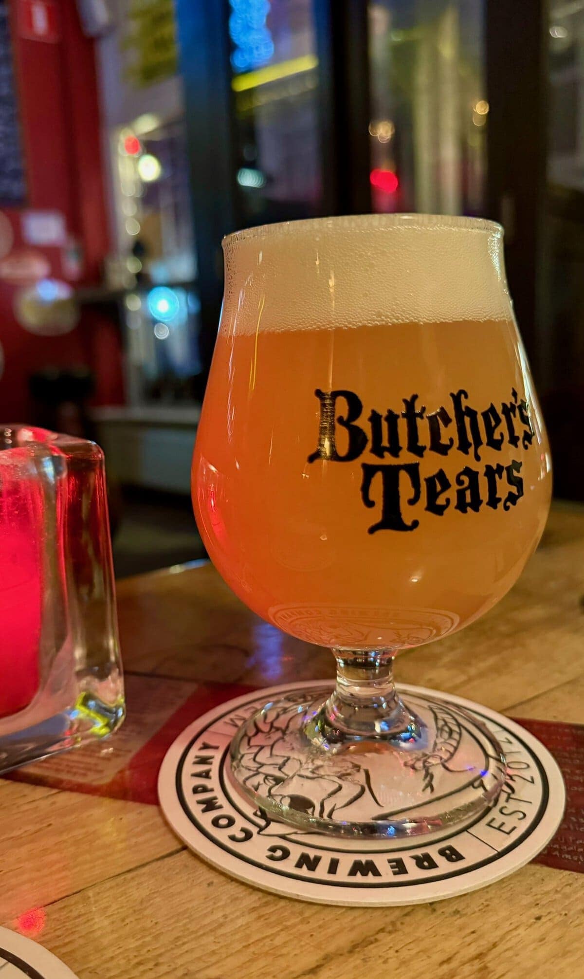 Cream-colored beer in a tulip-shaped glass with "Butcher's Tears" logo, served on a branded coaster at a cozy pub, highlighting European craft beer experience.